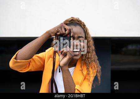 Glückliche afroamerikanische Frau, die fröhlich lacht und den Mund geöffnet hat, während sie mit der Fotokamera fotografiert Stockfoto