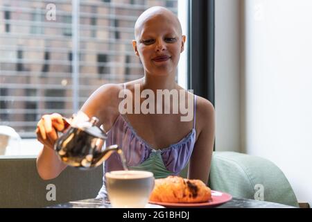 Positive junge, kahle Frau, die im Café auf dem Sofa sitzt, während sie ein Getränk von der Teekane in die Tasse auf dem Tisch gießt, mit Teller und Essen in der Nähe des Fensters Stockfoto