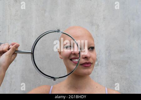 Doppelexposition von jungen Glatzenweibchen mit Alopezie-Autoimmunerkrankung, die einen runden Spiegel in der Nähe des Gesichts halten und unterschiedliche Reflexionen zeigen Stockfoto