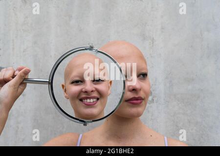 Doppelexposition von jungen Glatzenweibchen mit Alopezie-Autoimmunerkrankung, die einen runden Spiegel in der Nähe des Gesichts halten und unterschiedliche Reflexionen zeigen Stockfoto