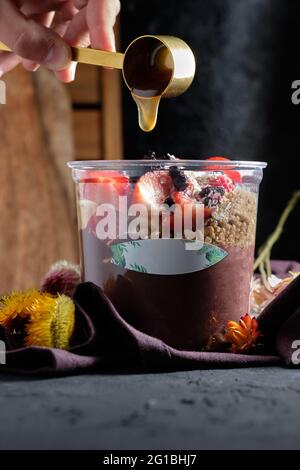 Crop anonyme Person gießt Honig in eine Schüssel appetitlich kalten Smoothie mit Müsli und frischen Beeren gekrönt Stockfoto