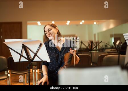 Fröhliche, professionelle Musikerin, die akustische Geige spielt und während der Probe im Studio Noten anschaut Stockfoto