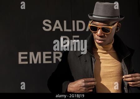 Trendiger, selbstbewusster afroamerikanischer Mann mit Hut und Sonnenbrille, der gegen eine graue Wand steht und wegschaut Stockfoto