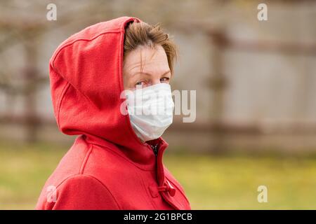 Eine ältere Frau mit braunem Haar und blauen Augen in einem roten Mantel und Kapuze in einer schützenden sicheren medizinischen Maske, Nahaufnahme Porträt. Stockfoto