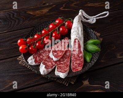 Auf einem schwarzen Teller auf einem rustikalen Holztisch werden katalanische Wurstscheiben und ein paar frische Kirschtomaten geschält. Traditionelle spanische, trocken gehäutete Schweinefleischsalami. Stockfoto