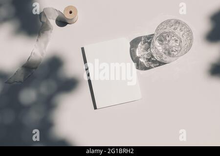 Sommer Hochzeit Schreibwaren Stillleben. Leere Grußkarte – Nachbau der Szene. Seidenband und Glas Wasser, Cocktail glitzert im Sonnenlicht. Beigefarbener Tisch Stockfoto