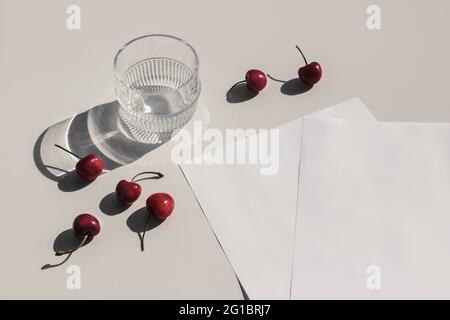 Sommer Essen Stillleben Zusammensetzung. Frische rote Kirschen Früchte auf beigefarbenem Tischhintergrund. Schreibwaren imitierte Szene. Leere Papierseiten. Glas Wasser Stockfoto