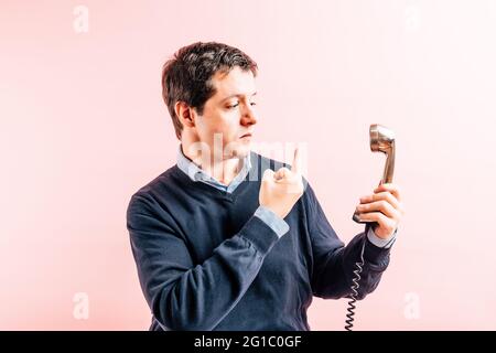 35-jähriger junger Erwachsener in blauem T-Shirt mit V-Ausschnitt und Pullover mit rosa Hintergrund, der einen Finger am schnurgebundenen Telefon hervorsticht. Concept ange Stockfoto