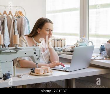 Junge fokussierte Frau in einem Werkstatt, die am Laptop arbeitet, während sie am Arbeitsplatz mit einer Nähmaschine sitzt und online Kleidungsdesign erstellt. Neue Kleid Kollektion hängen auf Kleiderständer stehen auf dem Hintergrund Stockfoto