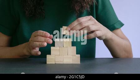 Frau Hand setzen einen quadratischen Holzblock auf Pyramidenstapel von Holzblöcken. Wachstums- oder Erfolgskonzept. Stockfoto