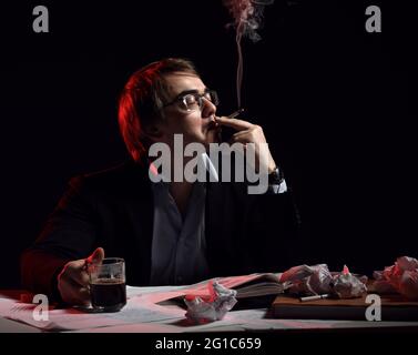 Der Erwachsene Mann in Hemd und Jacke sitzt hinter dem Schreibtisch, Schreibtisch mit Buch und Papieren, trinkt Tee, Kaffee und raucht Stockfoto