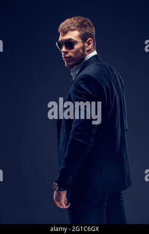 Seitenansicht eines Fashion bärtigen jungen ernsthaften Mannes in Sonnenbrille, Luxusanzug mit einer Hand in der Tasche Stockfoto
