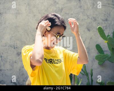 Sommerzeit. Lächeln Sie mit der Sonne. Schöne asiatische Frau kurze Haare tragen Sonnenbrille und gelbes Hemd setzen die weiße Plumeria Blume auf ihr Ohr Stockfoto