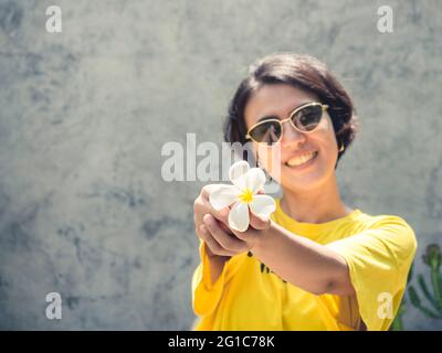 Sommerzeit. Lächeln Sie mit der Sonne. Die weiße Plumeria Blume auf der Hand der schönen asiatischen Frau kurze Haare tragen Sonnenbrille und gelbes Hemd auf Konz Stockfoto