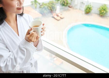 Beschnittenes Bild junge Frau im weißen Bademantel trinkt einen großen Becher Morgenkaffee, wenn sie am Hotelfenster steht und nach draußen schaut Stockfoto