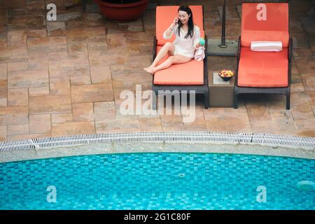 Fröhliche junge Frau, die auf der Chaiselounge am Pool sitzt, einen süßen Cocktail trinkt und mit einem Freund telefoniert Stockfoto