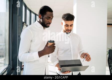 Zwei junge männliche Unternehmer diskutieren über ein neues Start-up, während sie mit einem Laptop mitten im Büro stehen Stockfoto