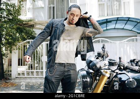 Schöner stilvoller Mann, der eine schwarze Lederjacke aufsetzt, wenn er bereit ist, auf dem Motorrad zu fahren Stockfoto