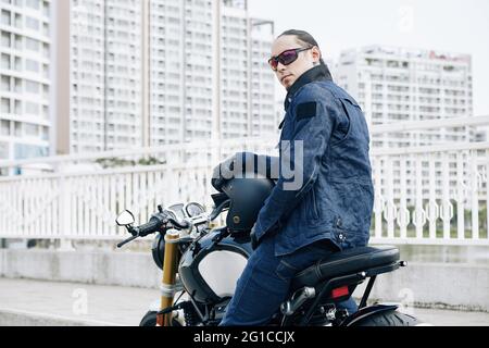 Ernsthafter Mann in Denim-Jacke und Sonnenbrille sitzt auf dem Motorrad und dreht sich zurück zur Kamera Stockfoto
