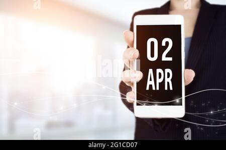 April. Tag 2 des Monats, Kalenderdatum. Weißes Smartphone mit Kalenderdatum in Geschäftsfrau Hand auf verschwommenem Hintergrund. Frühlingsmonat, Tag des Ye Stockfoto