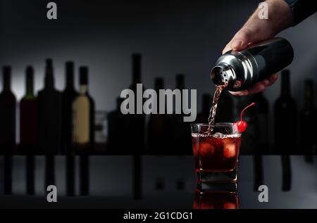 Cocktail „Manhattan“ auf schwarzem reflektierendem Hintergrund. Der Barkeeper gießt einen Cocktail aus einem Shaker in ein Glas mit Eis. Stockfoto