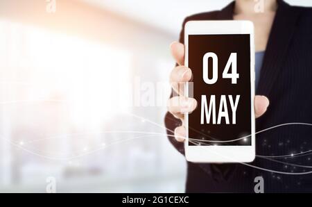 Mai. Tag 4 des Monats, Kalenderdatum. Weißes Smartphone mit Kalenderdatum in Geschäftsfrau Hand auf verschwommenem Hintergrund. Frühling Monat, Tag des Jahres Stockfoto