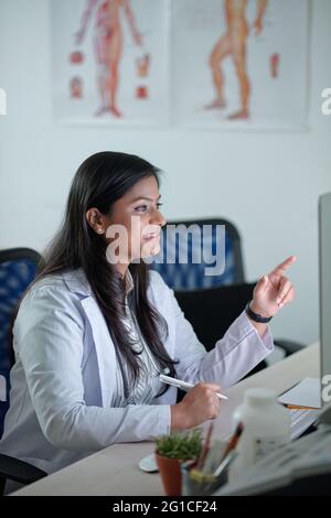 Lächelnd hübsche junge Ärztin, die Bericht oder medizinische Karte auf dem Computerbildschirm liest und sich Notizen in einem Dokument schreibt Stockfoto