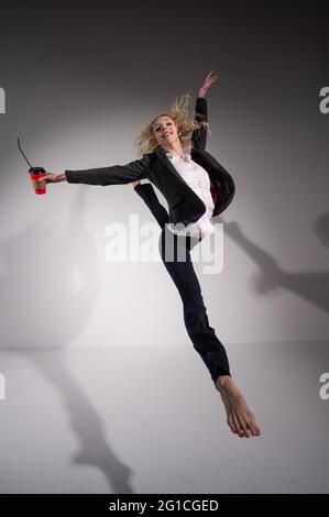 Anmutige Barfußballerina in einem Business-Anzug, die mit einem Glas Kaffee in den Händen auf weißem Hintergrund springt. Stockfoto