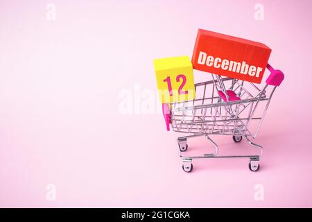 Dezember 12. Tag 12 des Monats, Kalenderdatum. Nahaufnahme Spielzeug-Metall-Warenkorb mit roten und gelben Box innen mit Kalender Datum auf rosa Hintergrund. Stockfoto