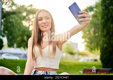 Glücklich lächelnde blonde Frau macht Selfie per Handy, Outdoor.Konzept der Urlaub Lebensstil. Schönes Mädchen mit Smartphone im Park.Sonniger Tag. Glückskonzept. Hochwertige Fotos Stockfoto