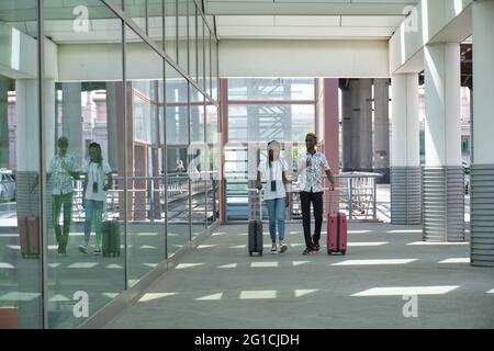 Ein junges schwarzes Paar mit Koffern, das in der Nähe des Bahnhofs entlang der Straße läuft und sich unterhält. Reisekonzept. Stockfoto