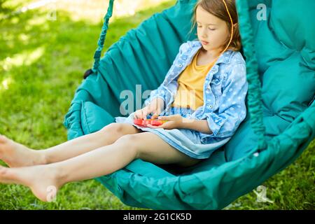 Ein kleines Mädchen, das auf einem hängenden Stuhl im Freien spielt, spielt Pop it, Kinderhände spielen Blasen eines Regenbogenentstreckungs-Spielzeugs, Fidget-Spielzeug im Hinterhof von Hou Stockfoto