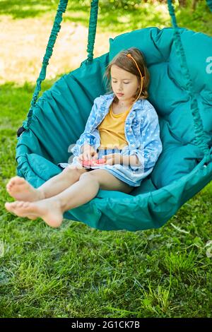 Ein kleines Mädchen, das auf einem hängenden Stuhl im Freien spielt, spielt Pop it, Kinderhände spielen Blasen eines Regenbogenentstreckungs-Spielzeugs, Fidget-Spielzeug im Hinterhof von Hou Stockfoto