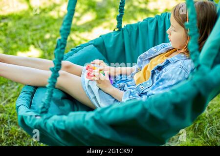 Ein kleines Mädchen, das auf einem hängenden Stuhl im Freien spielt, spielt Pop it, Kinderhände spielen Blasen eines Regenbogenentstreckungs-Spielzeugs, Fidget-Spielzeug im Hinterhof von Hou Stockfoto