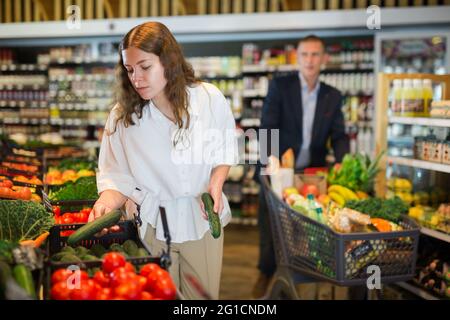 Das junge Mädchen mit einem Lebensmittelkarren wählt frisches Gemüse im Supermarkt aus Stockfoto