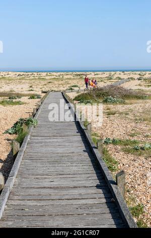 Ein Holzsteg über die Single zum Strand mit einer Familie, die zur Kamera in Dungeness, Kent, England, Großbritannien, geht. Stockfoto