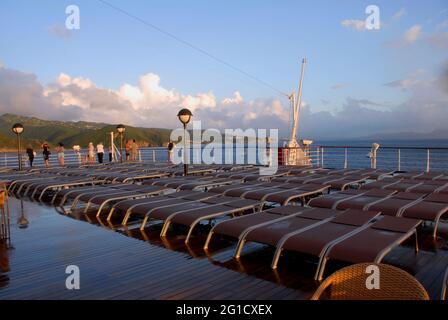 Auf dem Deck eines Kreuzfahrt-Liners mit leeren Sonnenliegen, nass nach Regen, Karibik und Passagiere, die auf der Schiene lehnen und die Aussicht bewundern Stockfoto