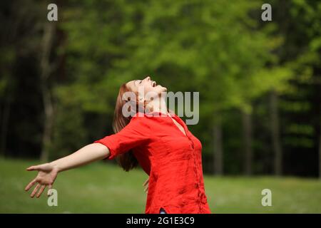 Aufgeregte Frau in Rot, die in einem Wald die Arme ausstreckt und in die Luft schreit Stockfoto