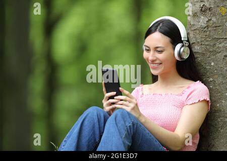 Glückliche asiatische Frau, die in einem Wald sitzt und Kopfhörer trägt und Musik hört Stockfoto