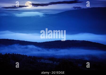 Leuchtender Nebel im ländlichen Tal bei Nacht. Wunderschöne Berglandschaft im Vollmondlicht. Blick vom Hügel Stockfoto