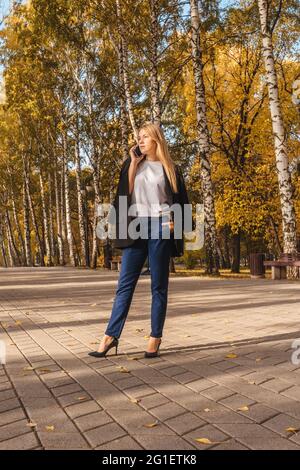 Geschäftsfrau in schwarzem Blazer, die im Park telefoniert, Ansicht von unten. Herbstpark, sonniger Tag. Stockfoto