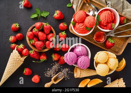 Haufen von Erdbeere und Orange, getrockneter Lavendel mit Tassen Eis und Zapfen. Lavendel, Erdbeere und Orangensorbet auf schwarzem Hintergrund. Stockfoto
