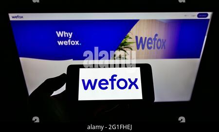 Person, die Mobiltelefon mit Logo der digitalen Versicherungsgesellschaft wefox Holding AG auf dem Bildschirm vor der Business-Webseite hält. Konzentrieren Sie sich auf die Telefonanzeige. Stockfoto