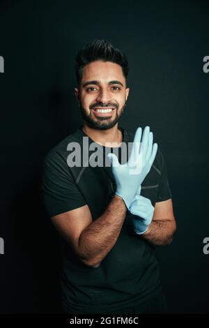 Porträt eines Mannes mit gemischter Rasse, der OP-Handschuhe trägt und mit glänzenden Zähnen vor schwarzem Hintergrund lächelt Stockfoto