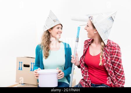 Lächelnde Freundinnen tragen einen Papierhut, der den Farbkübel und die Rolle vor weißem Hintergrund hält Stockfoto