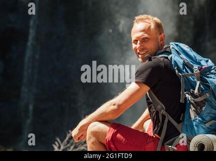 Mit freundlichen Grüßen lächelnder Mann Porträt mit Rucksack in aktiven Trekking-Kleidung in der Nähe von Berg Fluss Wasserfall lächelnd und genießen Sie die Natur gekleidet. Stockfoto