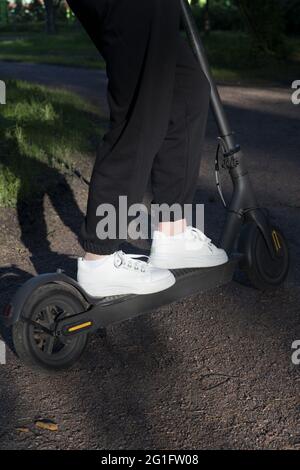 Die Beine der jungen Frau sind mit schwarzen Jogginghosen und weißen Sneakers auf einem Elektroroller gekleidet Stockfoto