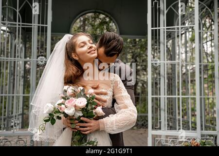 Junger Mann küsst Hals der glücklichen Braut hält Hochzeit Strauß im Park Stockfoto