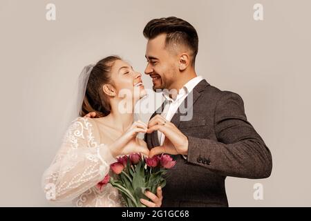 Fröhliche Brautpaar lächeln einander an, während sie ein Herzsymbol mit Händen auf Grau zeigen Stockfoto