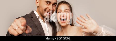 Fröhliche Jungvermählte winken mit der Hand und zeigen mit Finger isoliert auf grauem Banner Stockfoto